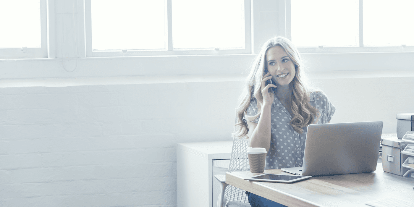 person sitting at a table at the office with a laptop and cup of coffee, speaking on the phone
