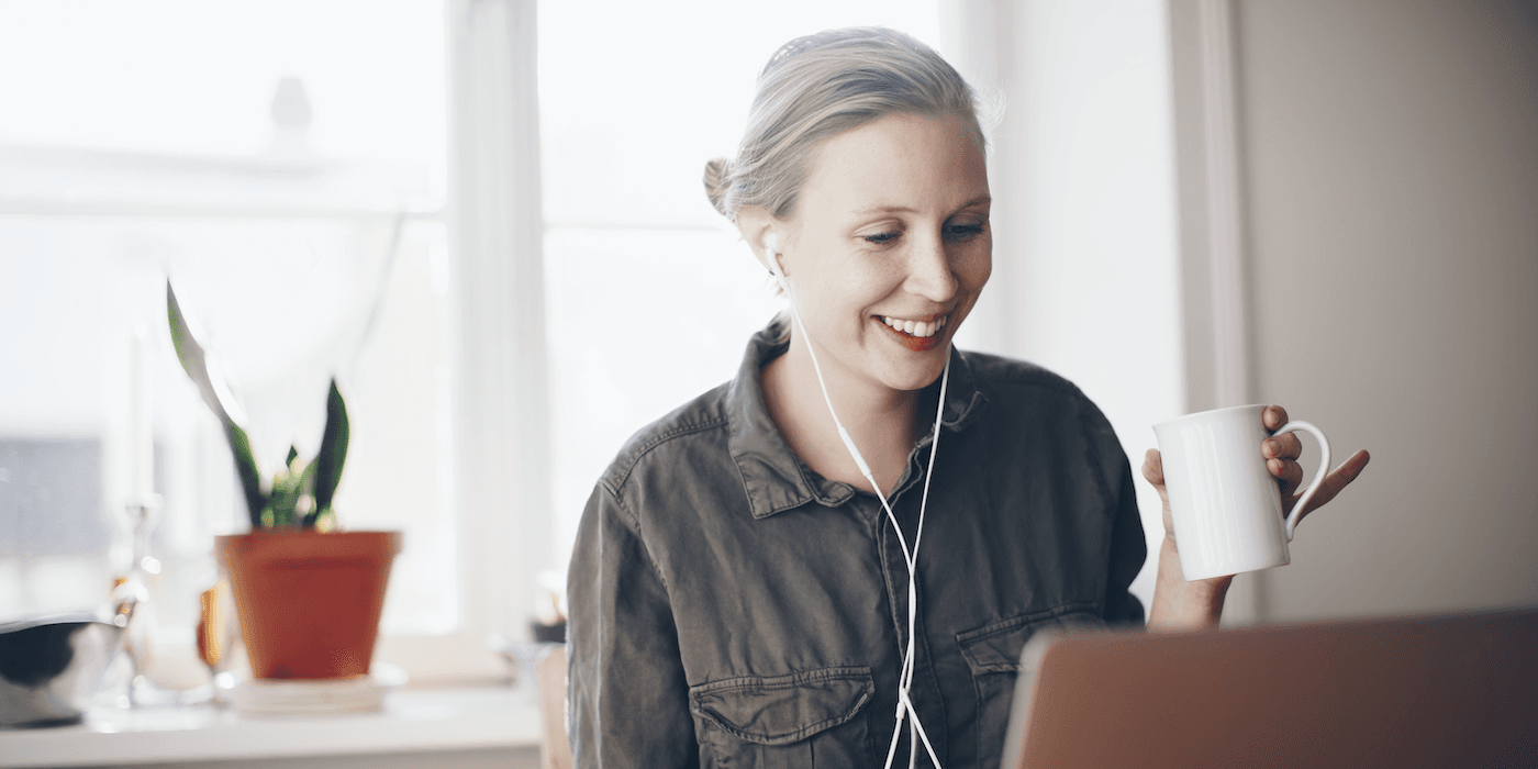person sitting at home in front of laptop, wearing headphones, smiling, and holding a mug