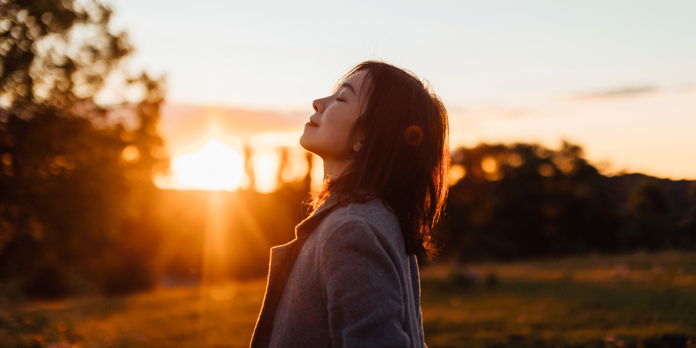 person standing outdoors in a light jacket during sunset or sunrise, eyes closed, head tilted up