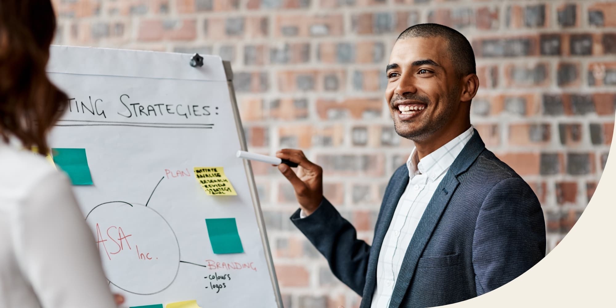 smiling person standing in front of brick wall holding a marker and placing sticky notes on large white pad on stand that says “strategies”; coworker facing away from camera in foreground