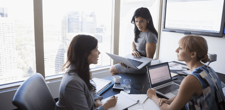 three people sitting at an office table with laptops and notepads